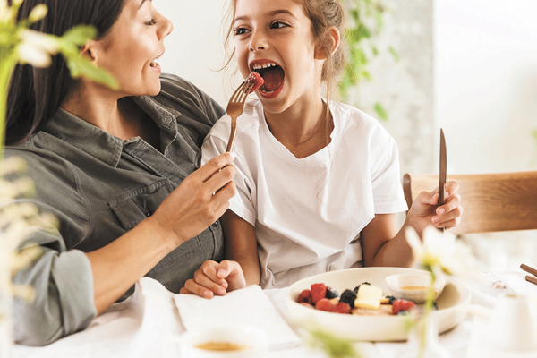 Mom sitting at dining room table feeding kid on her lap piece of fruit from fork