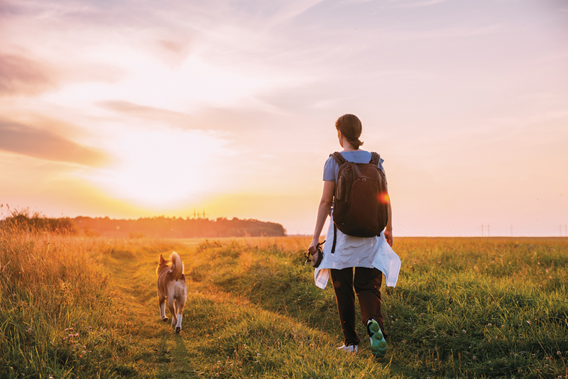 Person wearing backpack walking outside along path at sunset with dog at side
