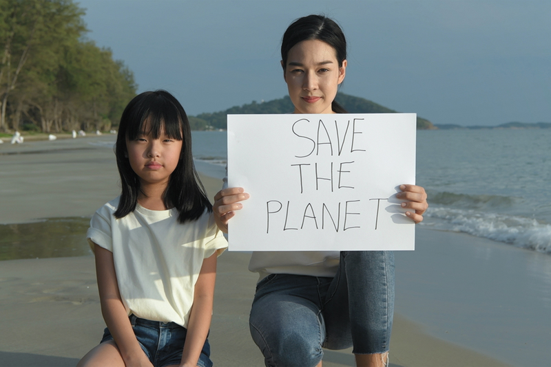 Parent with child kneeling on the beach shoreline holding sign reading "Save the Planet"