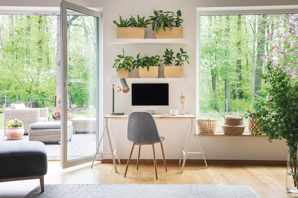 Interior of home with computer monitor on desk, chair, floating shelves with indoor plants, large windows showing trees and nature, outdoor back patio with furniture